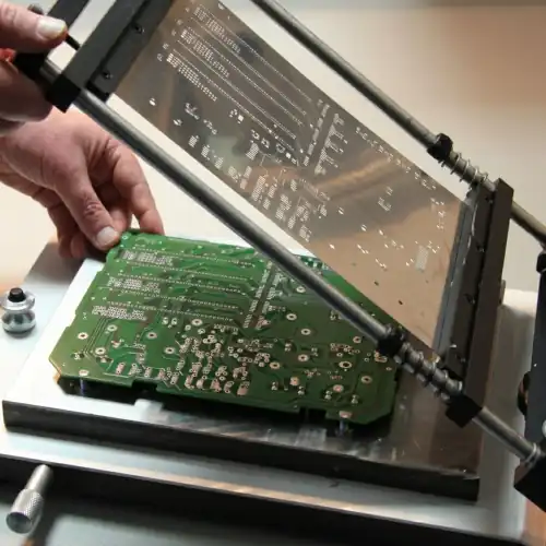 Hand holding green PCB prototype with metal testing fixture or assembly jig in background, demonstrating prototype inspection and assembly workflow.