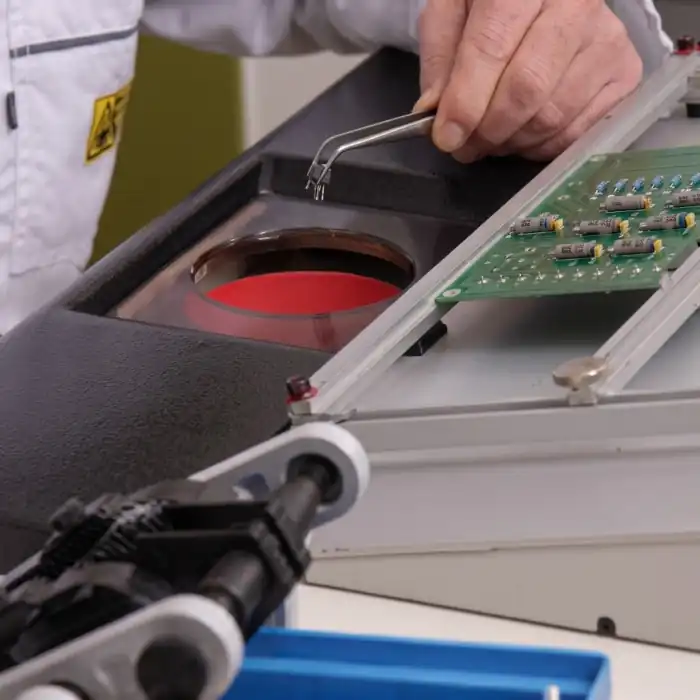 Technician performing precision electronics THD assembly or inspection with tweezers over a thermal inspection chamber containing circuit components.